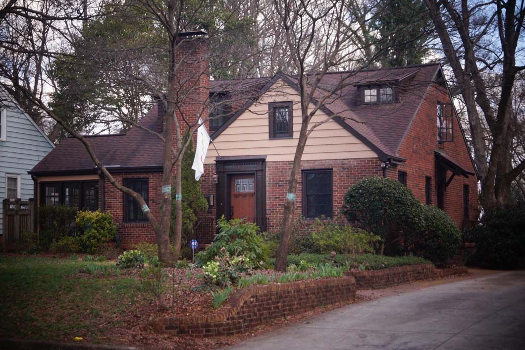 Red brick house with a brown roof, chimney, and dormer windows, in a setting with trees and a small garden.