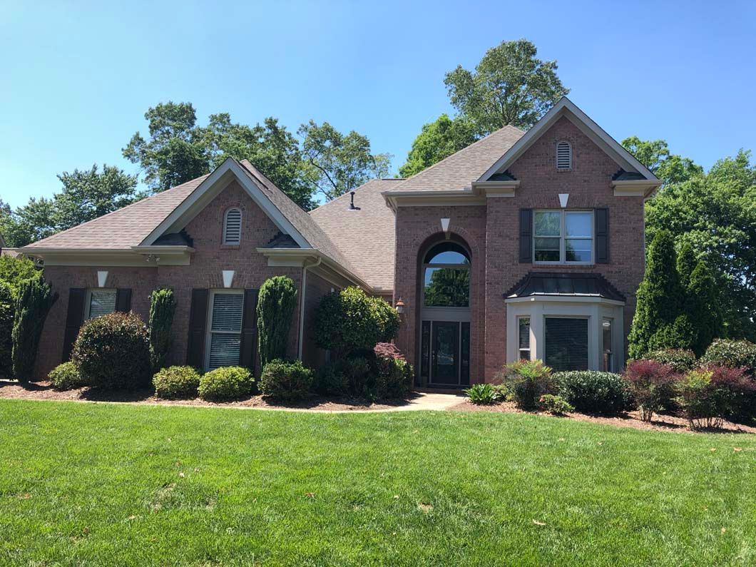 Brick house with a brown roof, green lawn, and shrubbery under a blue sky.