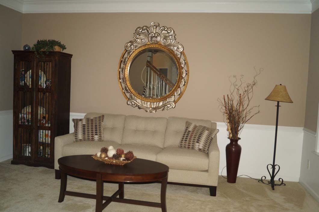 Living room with tan walls, white wainscoting, beige sofa, round mirror, dark wood cabinet, coffee table, lamp, and vase.