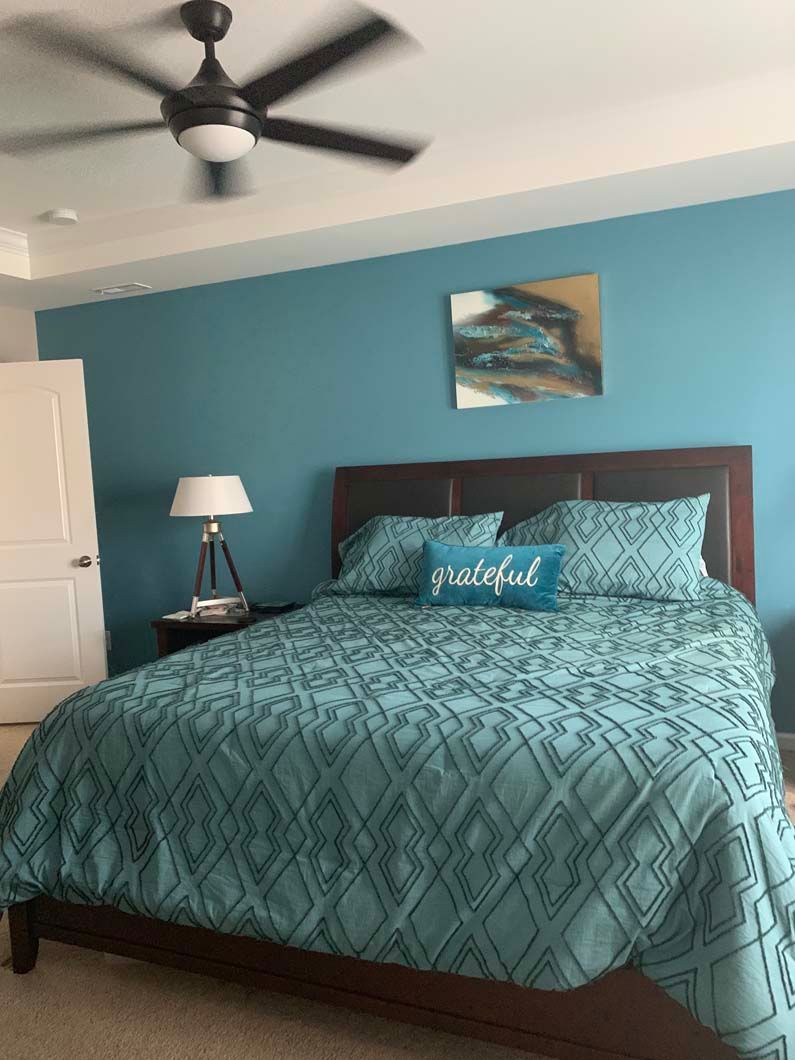 Bedroom with teal accent wall, bed, artwork, lamp, and ceiling fan.