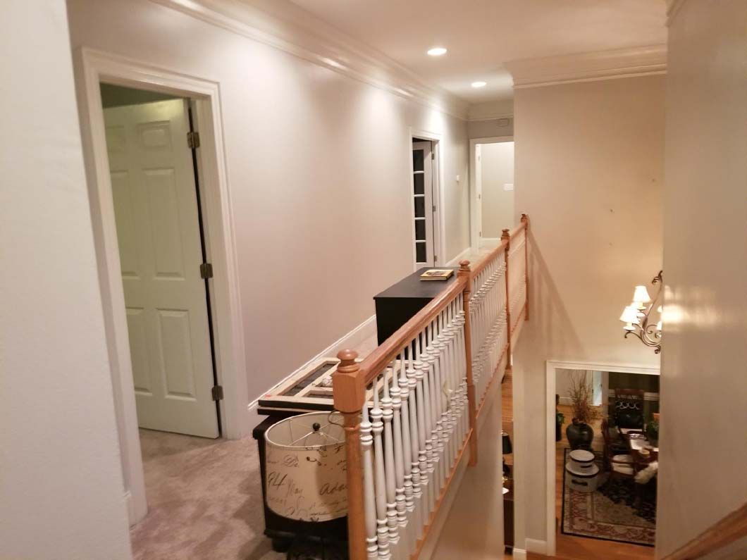 Hallway with a staircase, doors, and a decorative railing. Beige walls, wood accents, and overhead lighting.