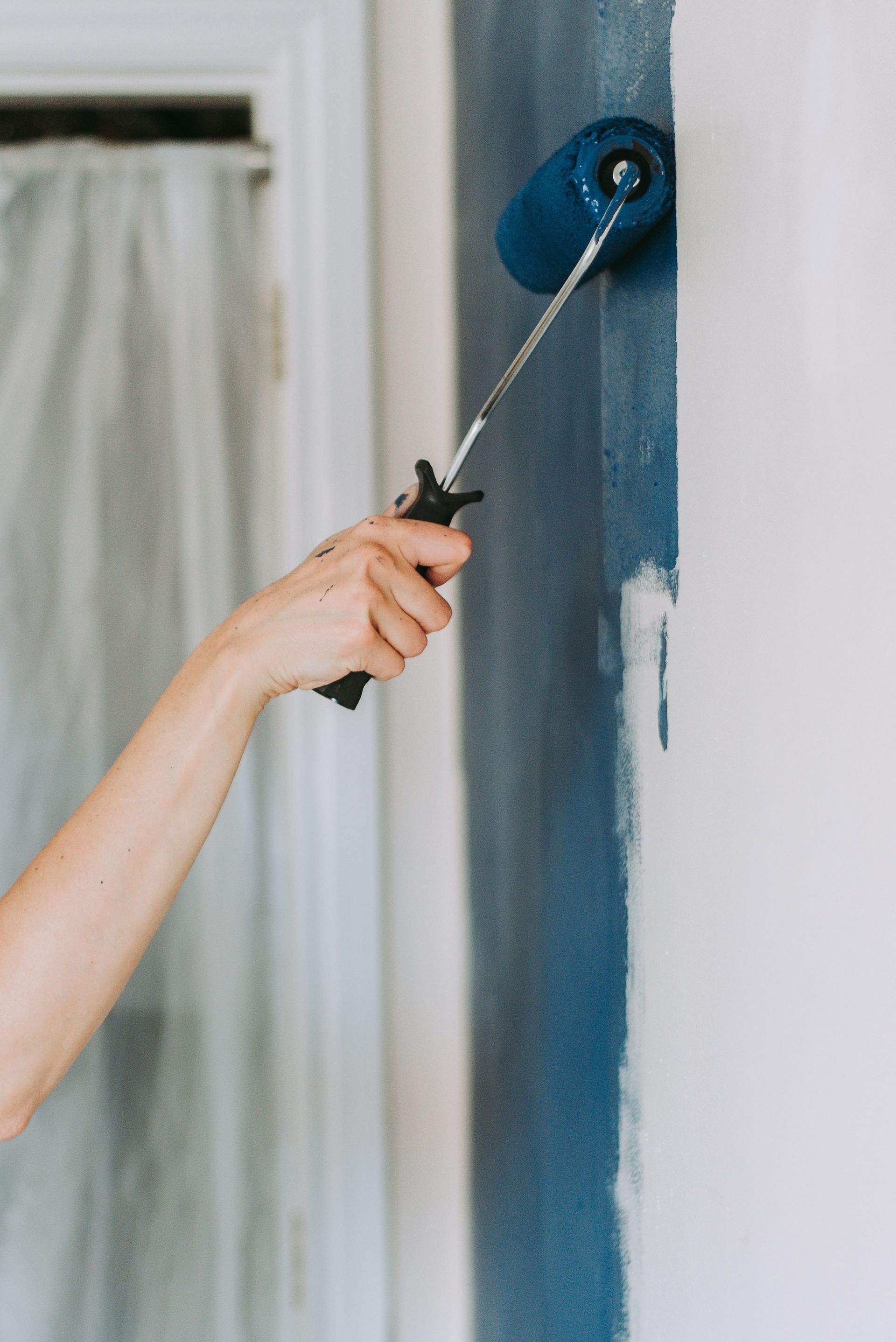 A person paints a wall blue with a roller; interior setting, white wall, window.