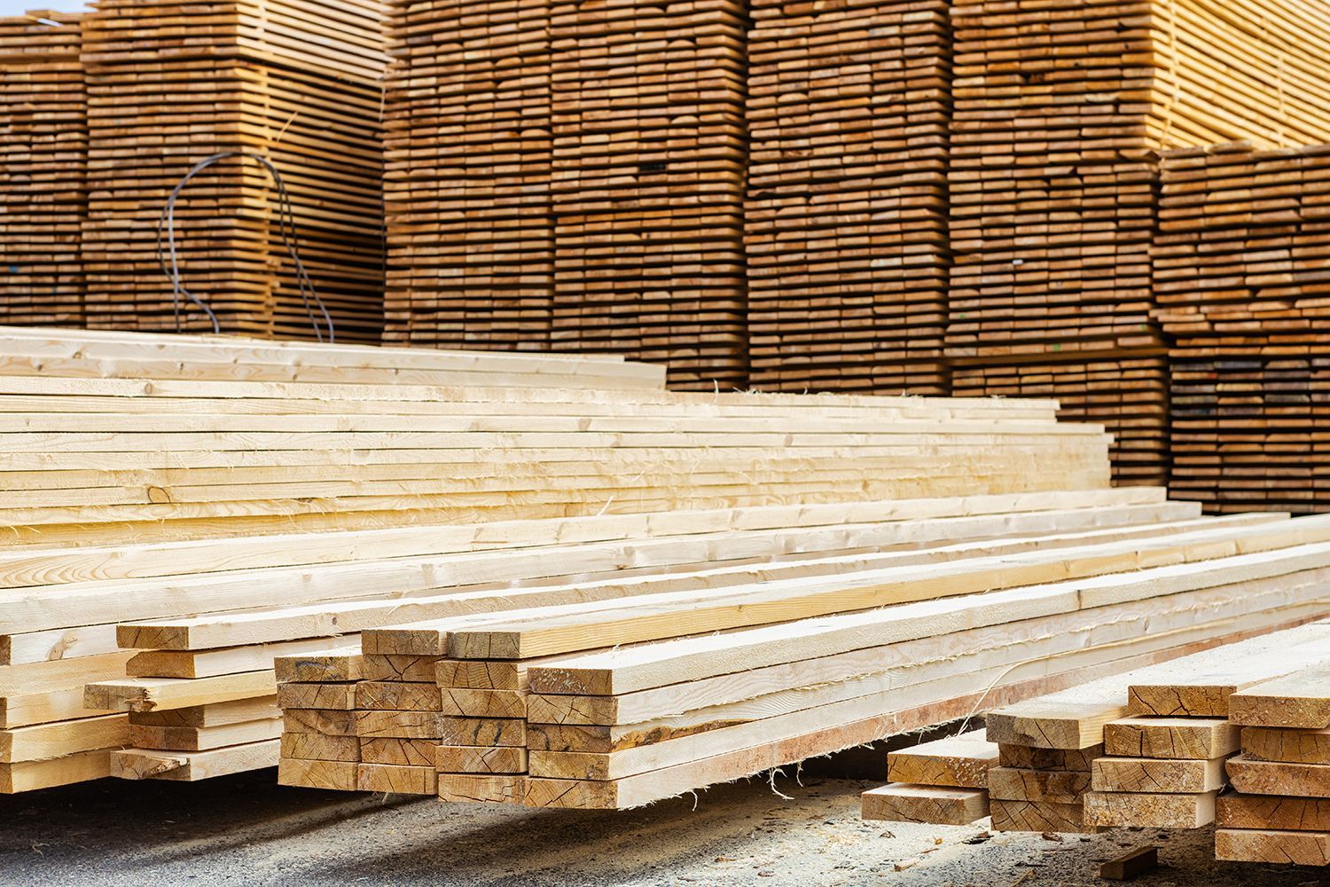 Stacks of lumber boards in a warehouse yard, with long white planks in the foreground and brown stacked wood behind.
