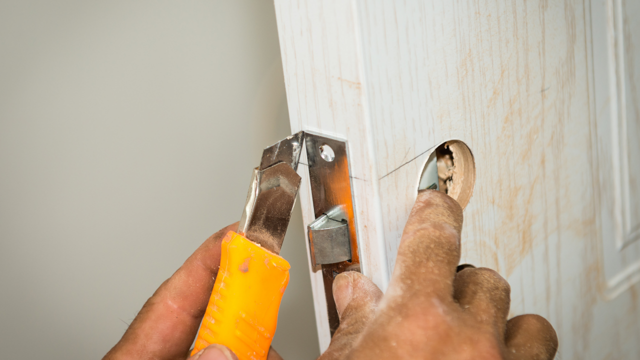 Hand using a hammer and chisel to remove a door latch from a white wooden door