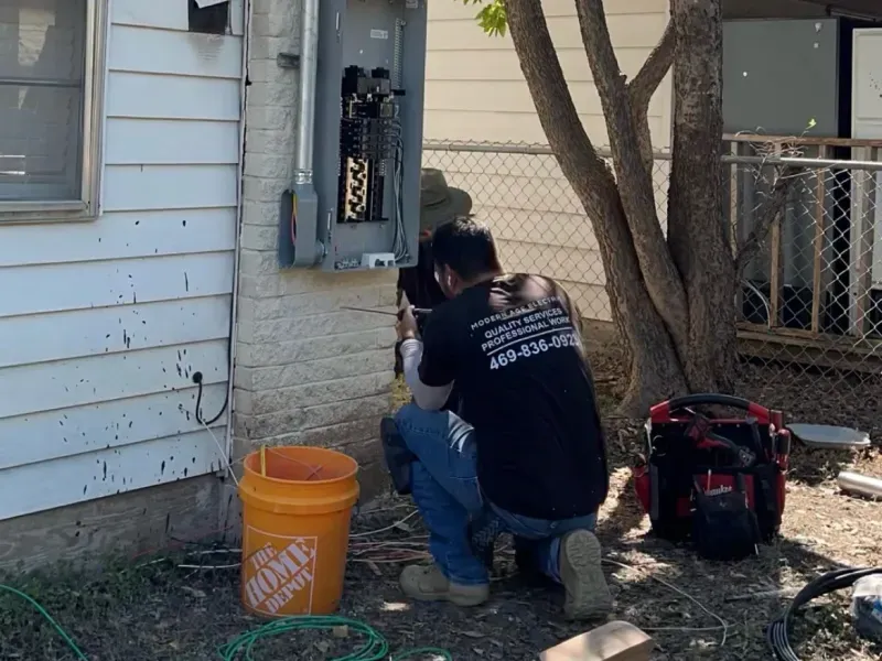 Person kneeling by an outdoor electrical panel beside a house, with orange bucket and tools nearby.