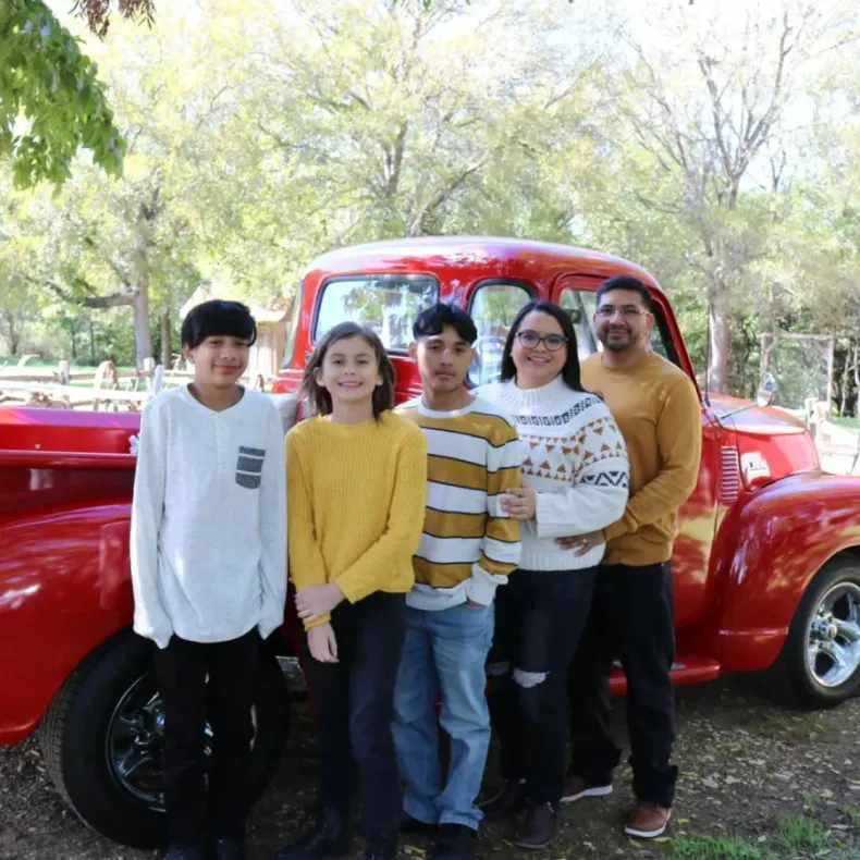 Five people posing in front of a bright red vintage car in a park, with trees in the background.