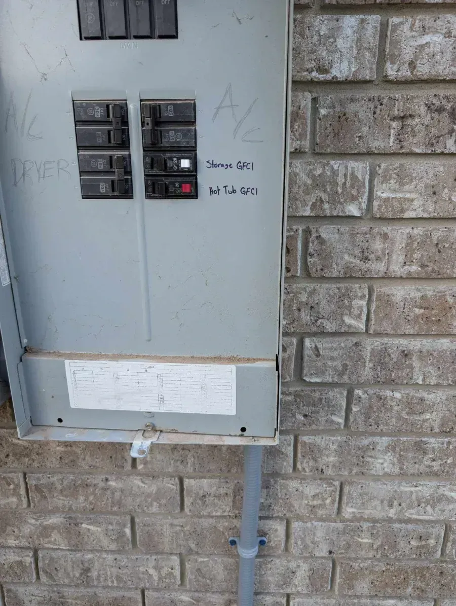 Gray electrical breaker panel mounted on a brick wall with handwritten labels and a conduit pipe below.