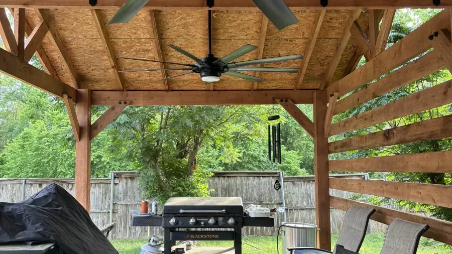 Covered backyard patio with wooden rafters, ceiling fan, grill, chairs, and fenced lawn beyond