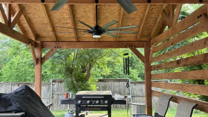 Covered backyard patio with wooden rafters, ceiling fan, grill, chairs, and fenced lawn beyond