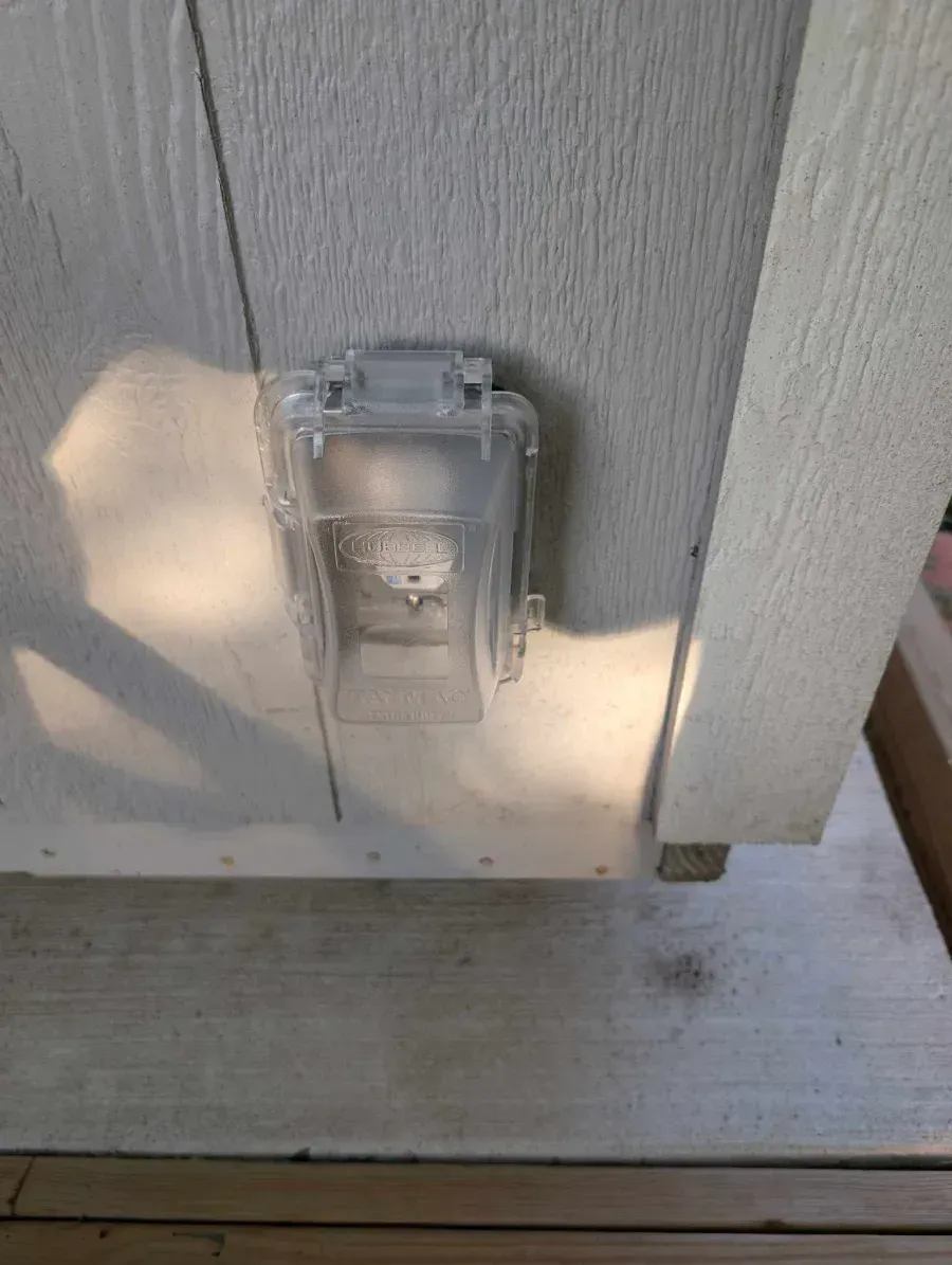 Clear plastic outdoor electrical outlet cover on a gray siding wall beside a concrete step.