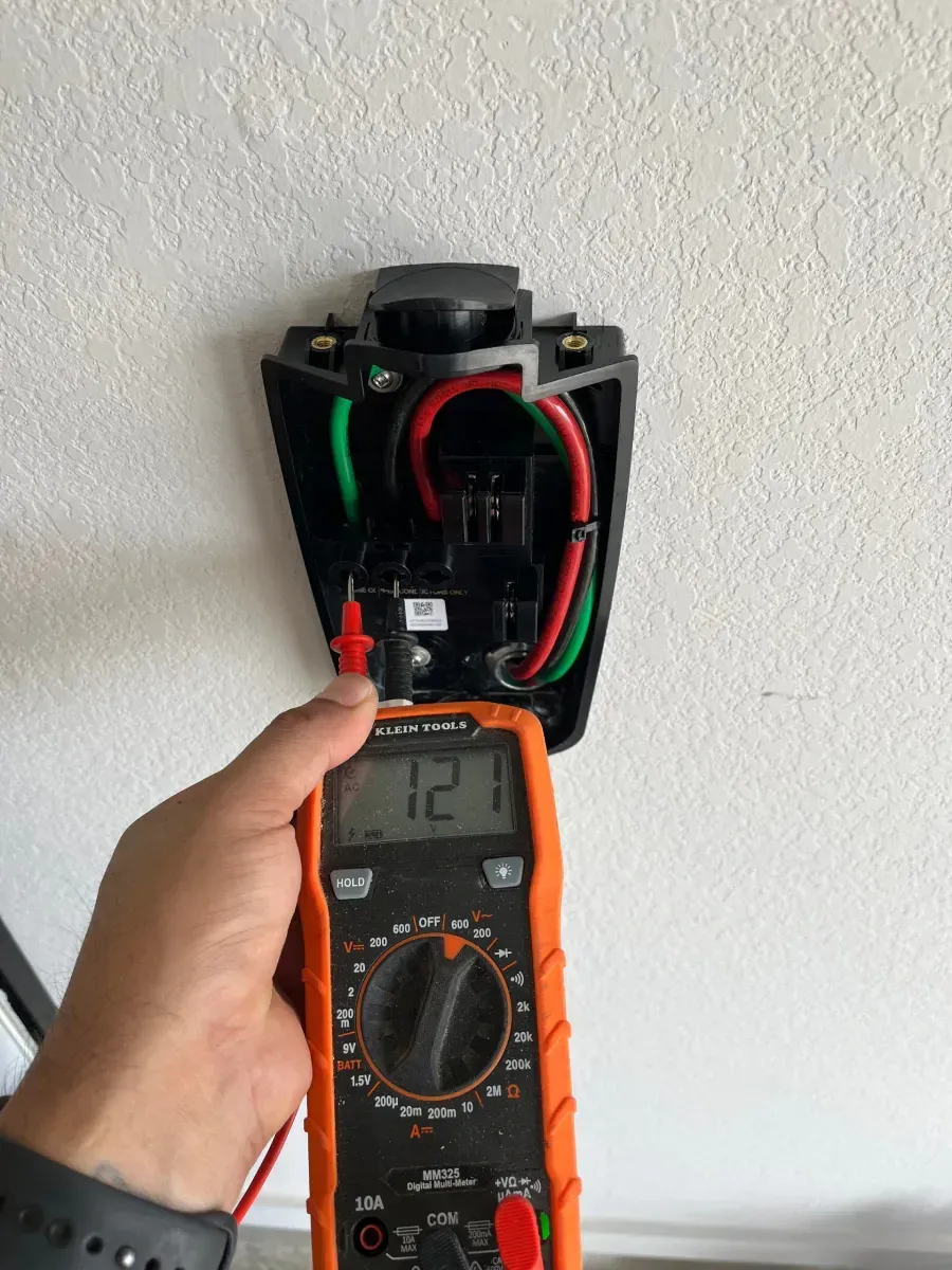 Hand holding a multimeter to an open electrical outlet box with red and green wires exposed