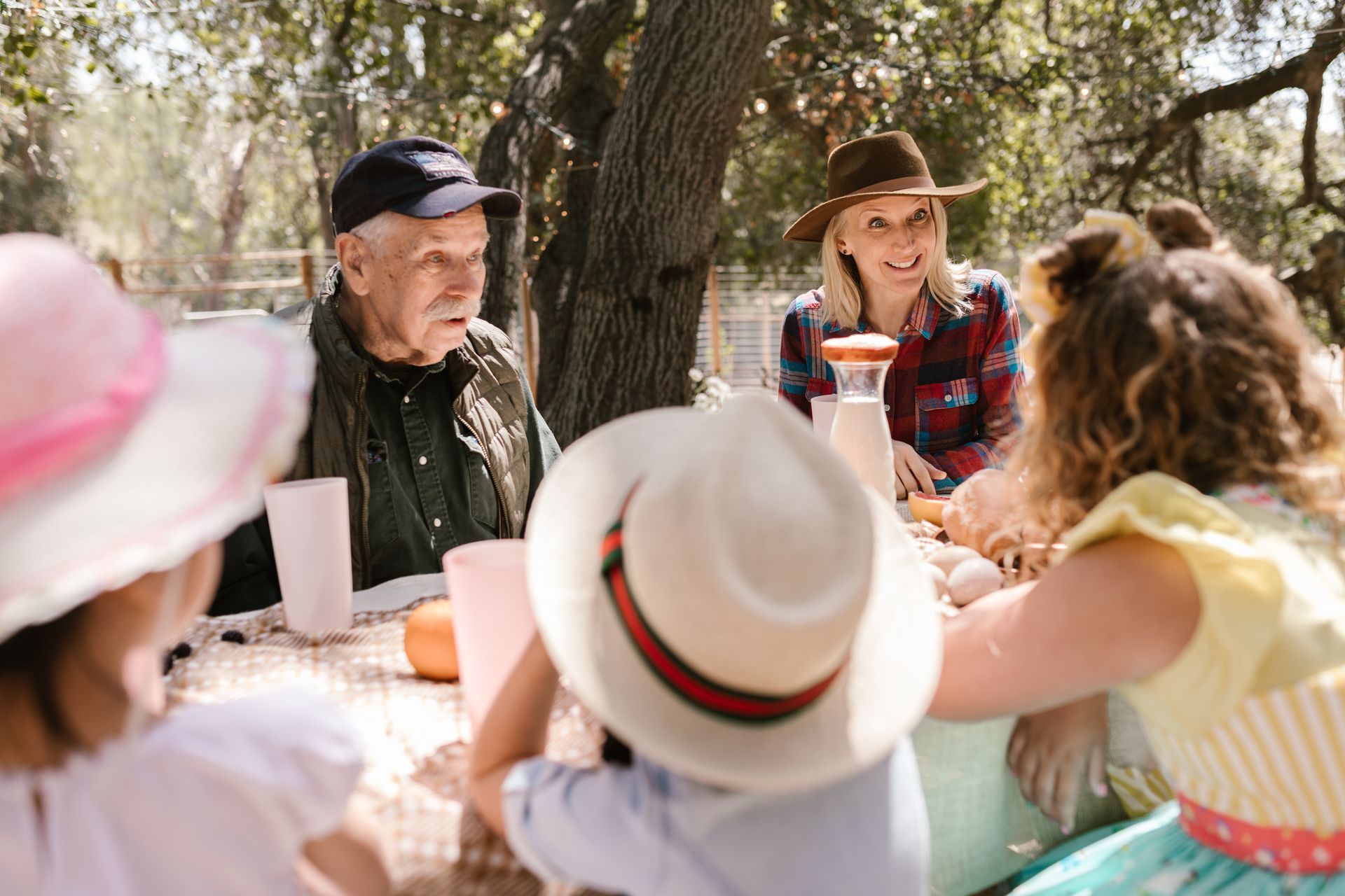 People gathered around a picnic table outdoors under trees, talking and wearing colorful hats.