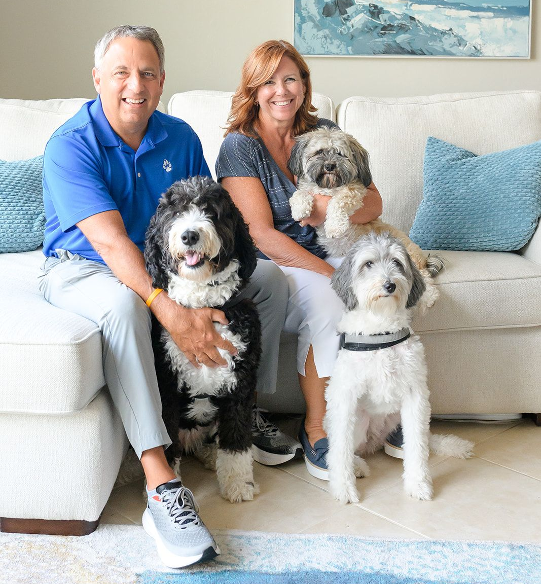 Couple seated on a couch with three fluffy dogs in a bright living room