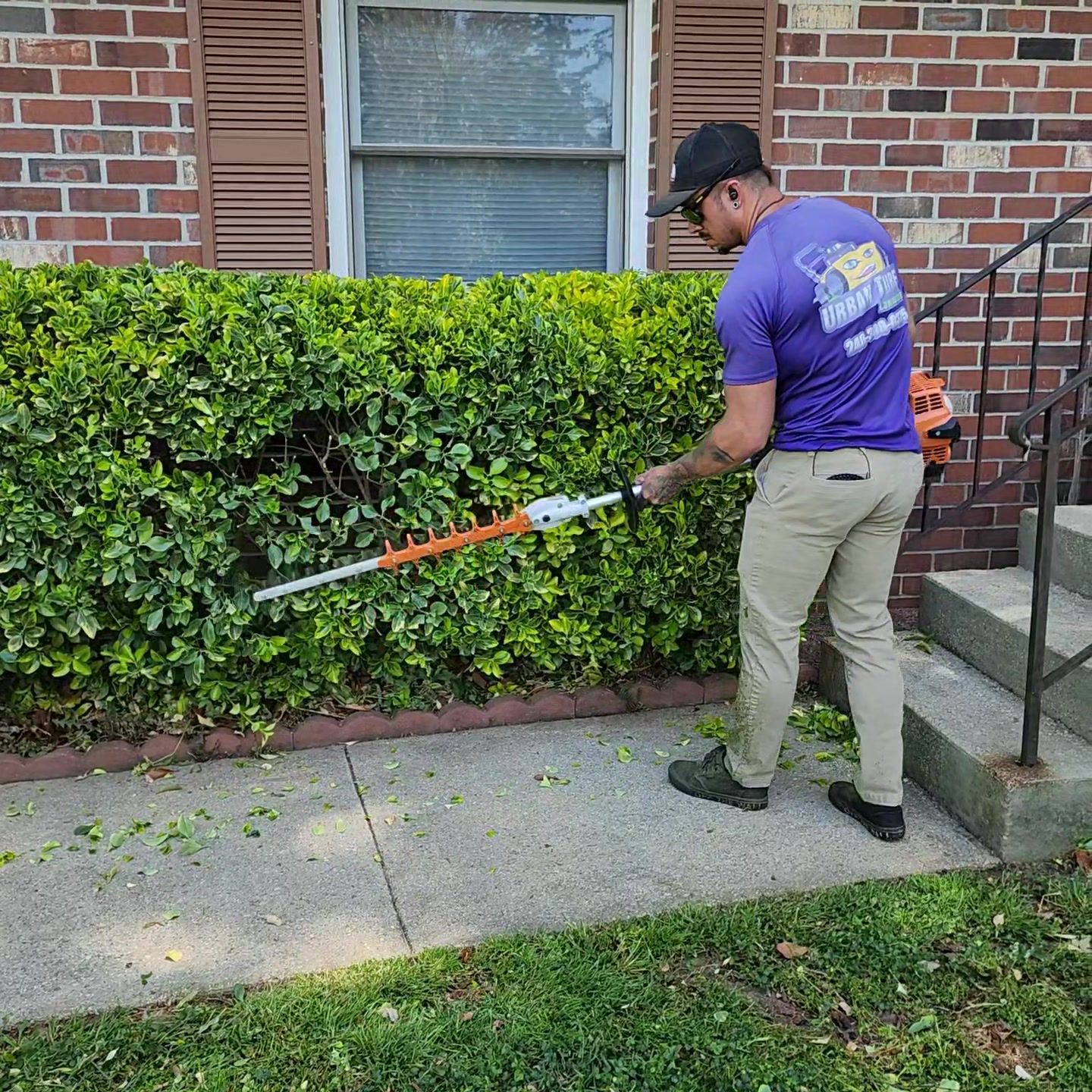 A person trims a hedge with a hedge trimmer near a house.