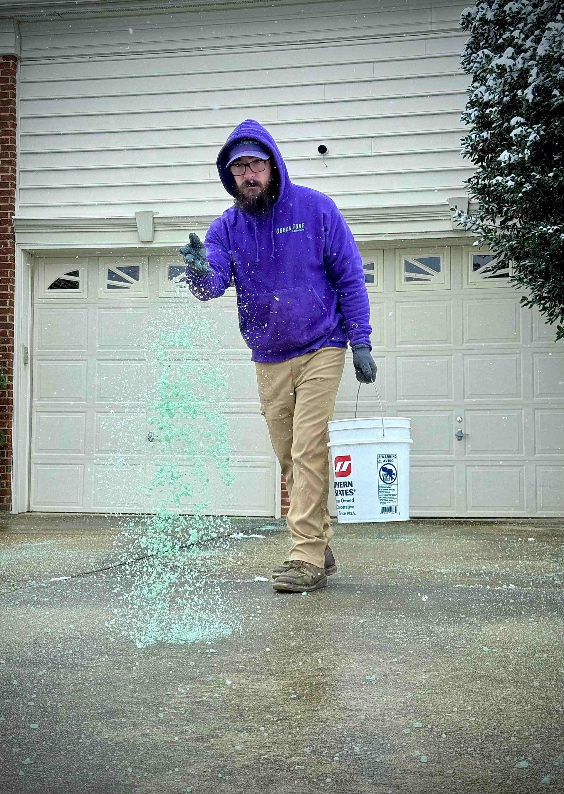 Man in purple hoodie spreading salt on a snow-covered driveway in front of a garage.