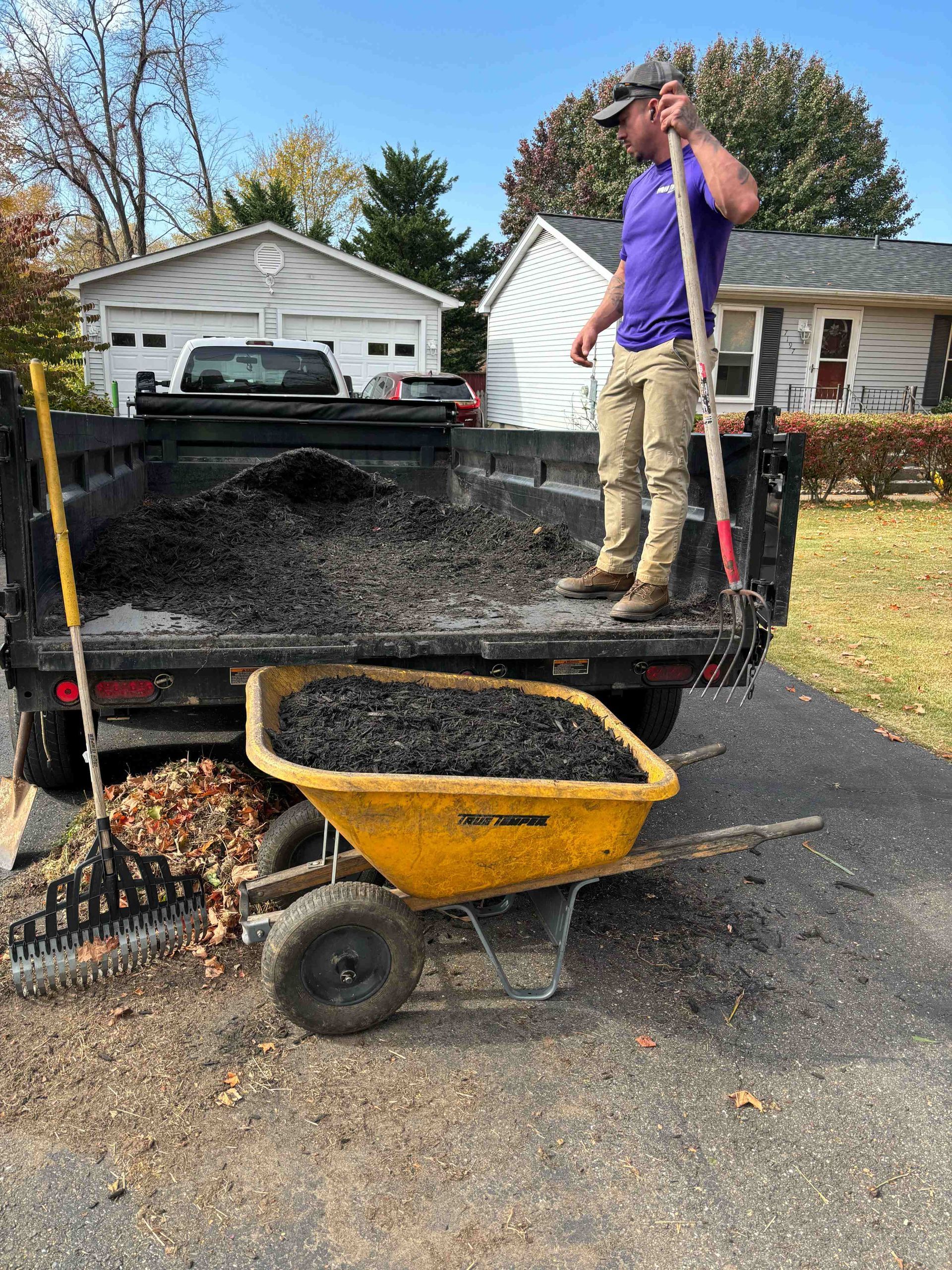 A person rakes debris into a truck bed, using a yellow wheelbarrow. Houses and a tree are in the background.