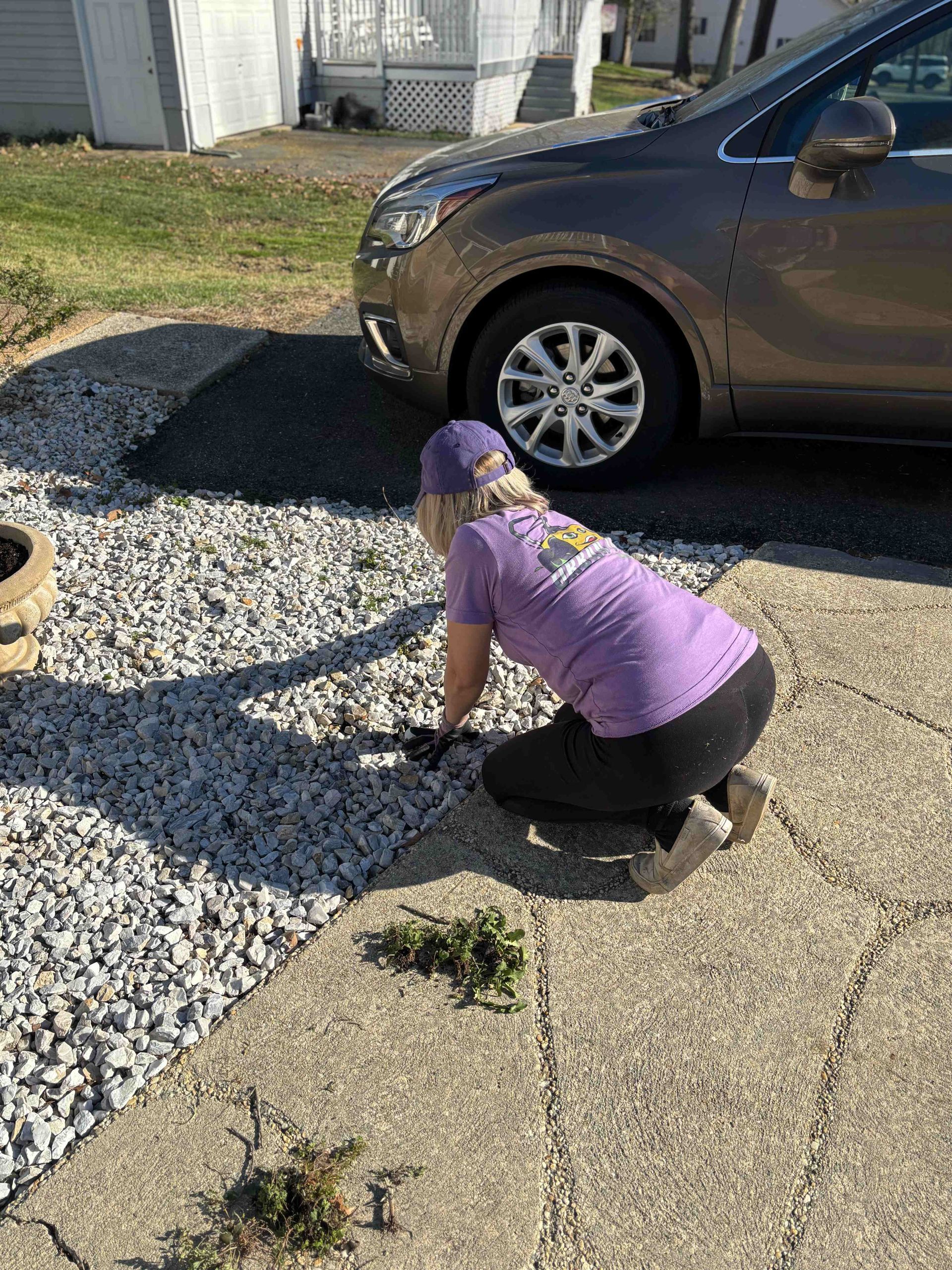 Woman kneeling, weeding between a concrete walkway and gravel driveway next to a parked car.