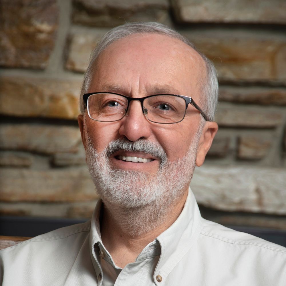 A man with glasses and a beard is smiling in front of a stone wall.