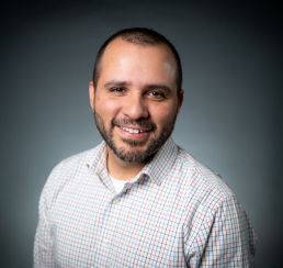 A man in a suit and white shirt is standing in front of a white background.