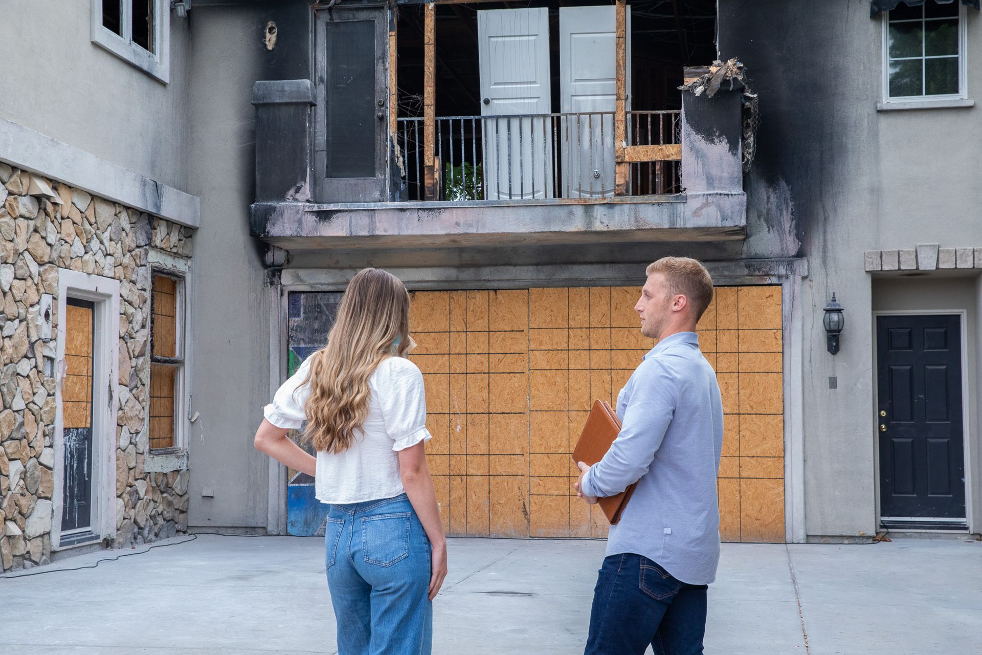 A couple surveys fire-damaged home. Man holds clipboard, woman stands with hand on hip.