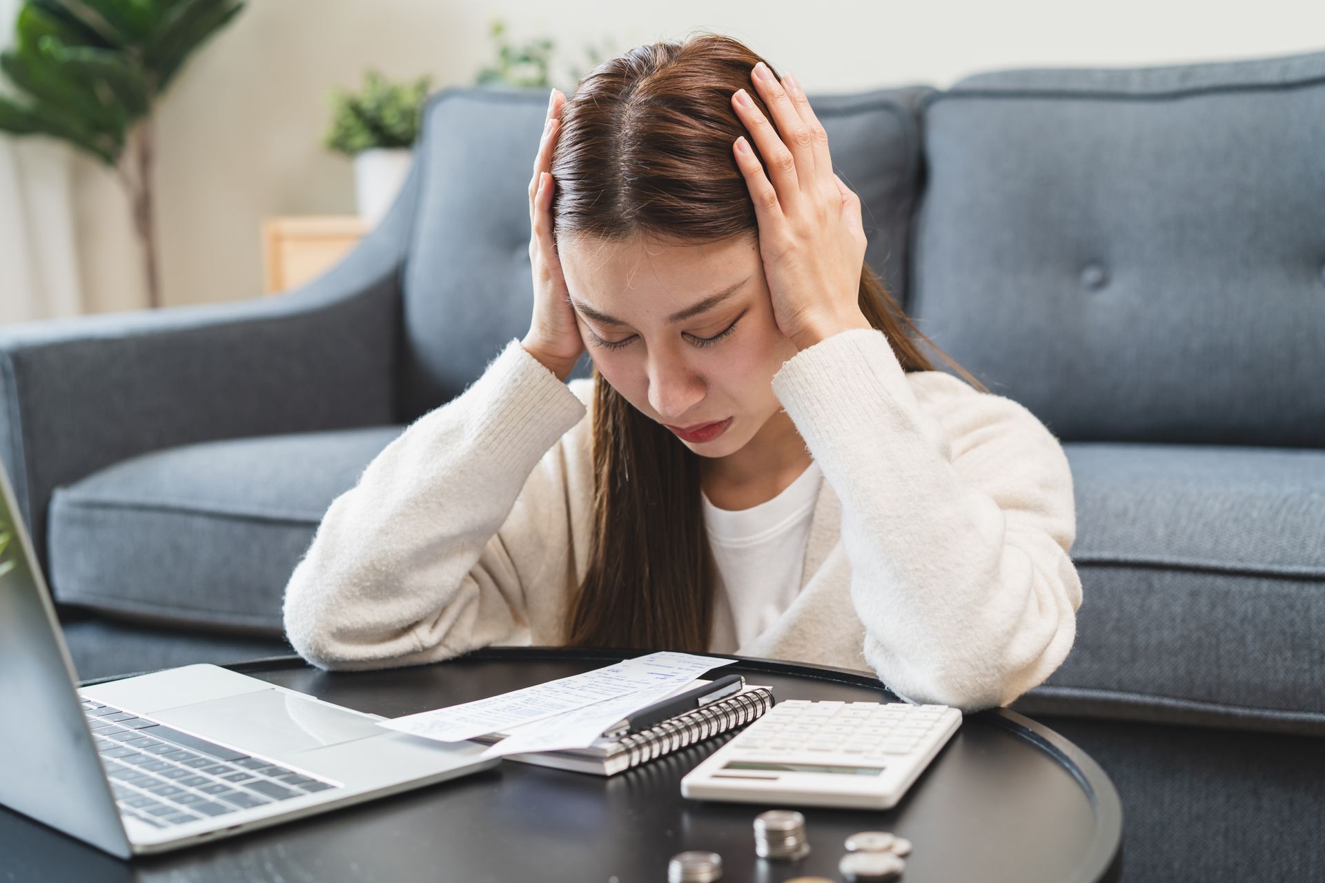 Woman with hands on head, stressed by finances, surrounded by bills, laptop, and calculator.
