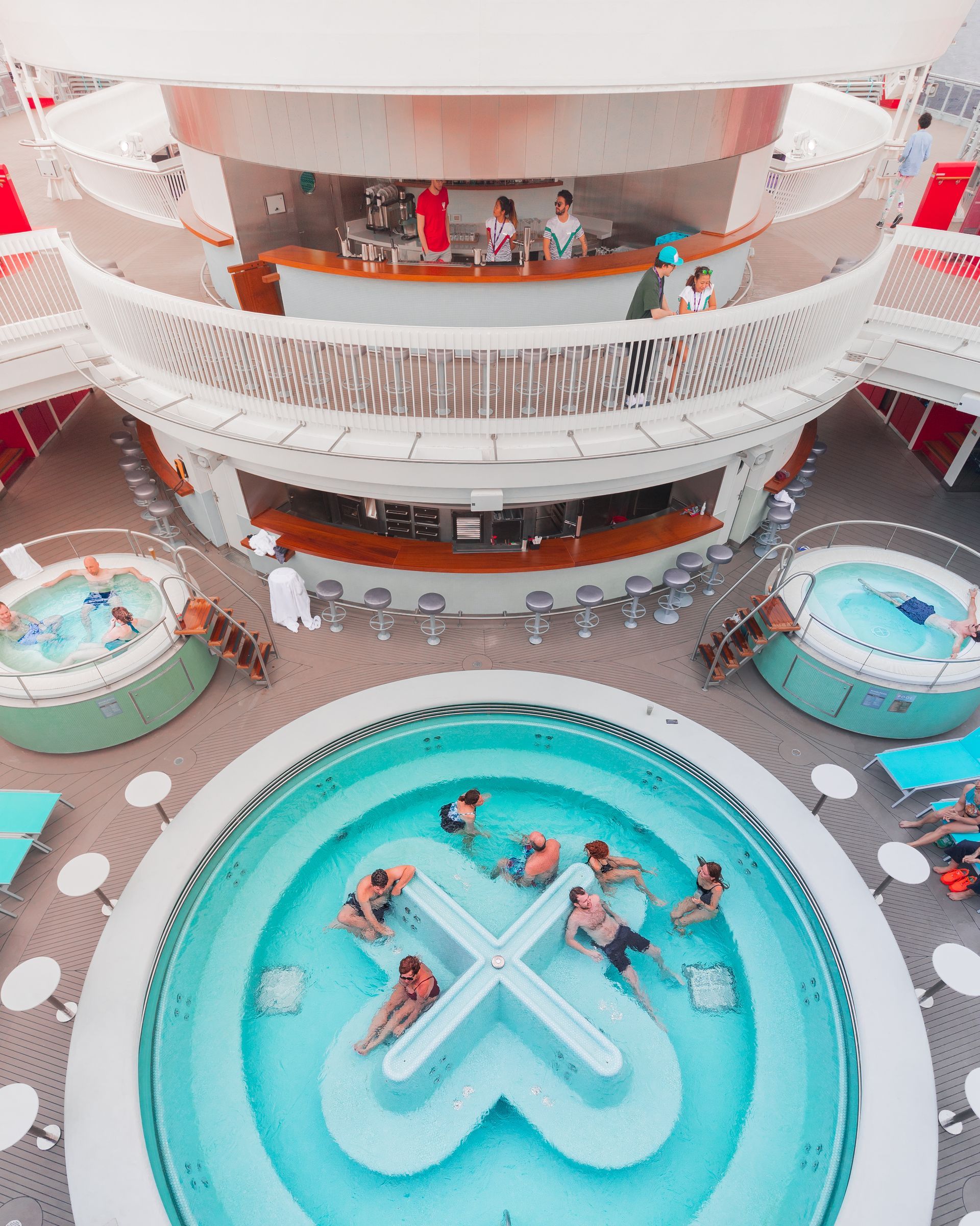 Cruise ship pool deck with people lounging around a heart-shaped turquoise pool and red-white balconies