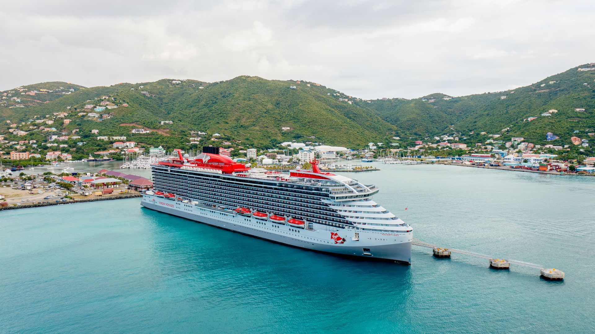 Large red-and-white cruise ship in a turquoise harbor beside a green hillside town