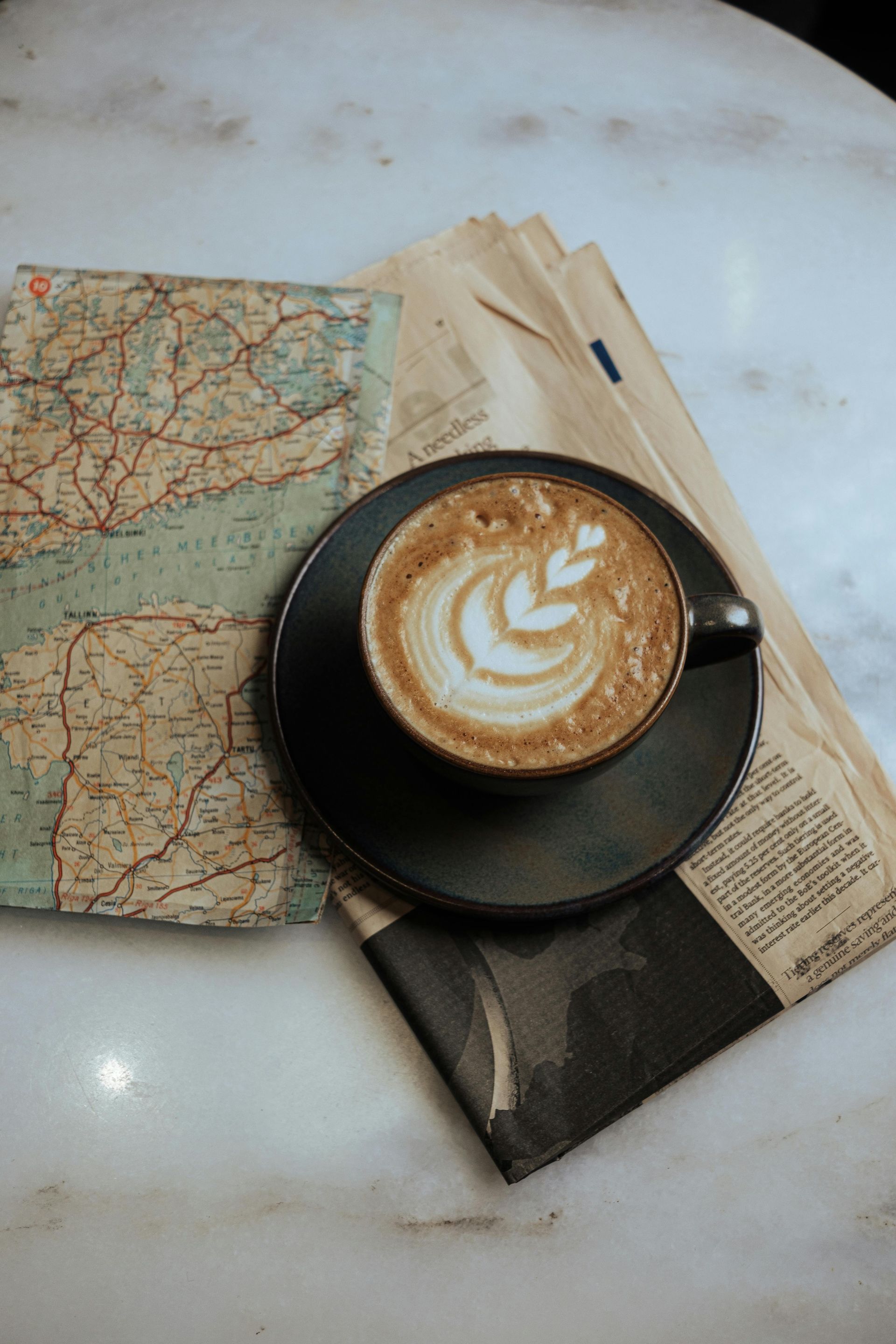 Latte with leaf art sits on black saucer atop newspaper and map on marble table.