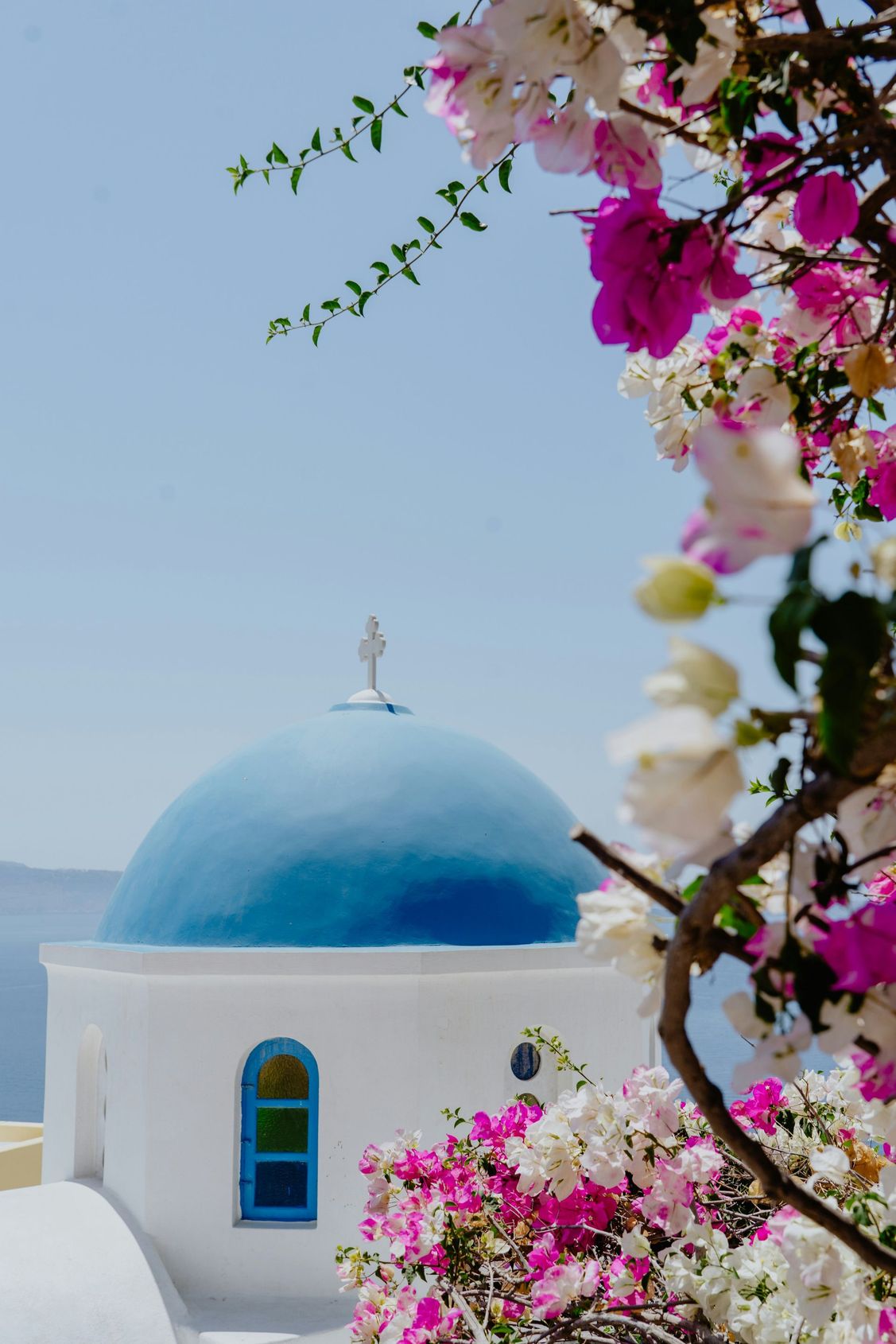 Stone steps descend towards the blue sea, lined with white buildings, bougainvillea, and a small tree.