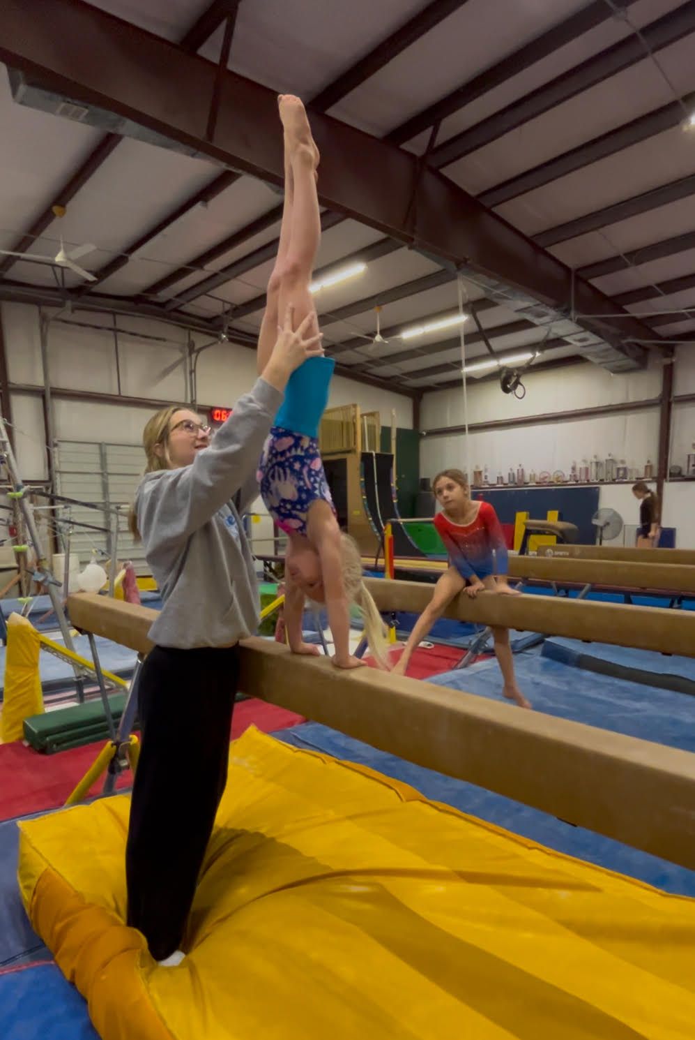 A girl is doing a handstand on a balance beam in a gym.