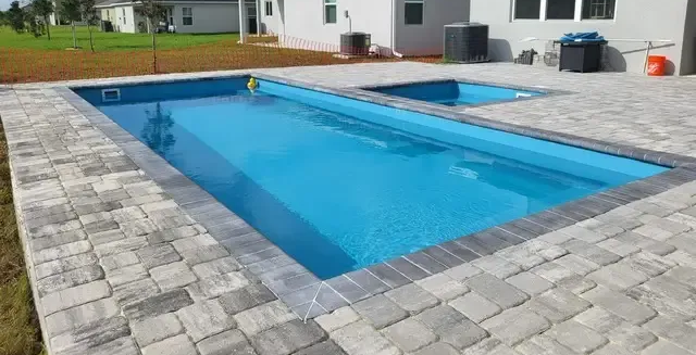 Inground swimming pool with wide paver decking and lounge chairs beside a coastal home under an overcast sky