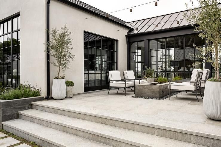 Outdoor patio with seating, fire pit, potted trees, and large black-framed windows.