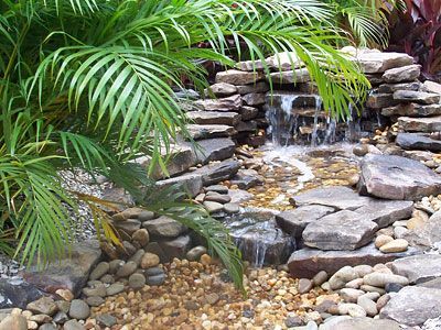 Small stone waterfall in a garden, with lush green plants surrounding the water feature and pebbles.