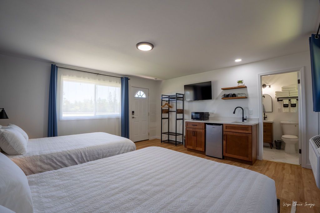 Bedroom with two beds, small kitchen area, and bathroom. White walls, wood floors, and blue curtains.