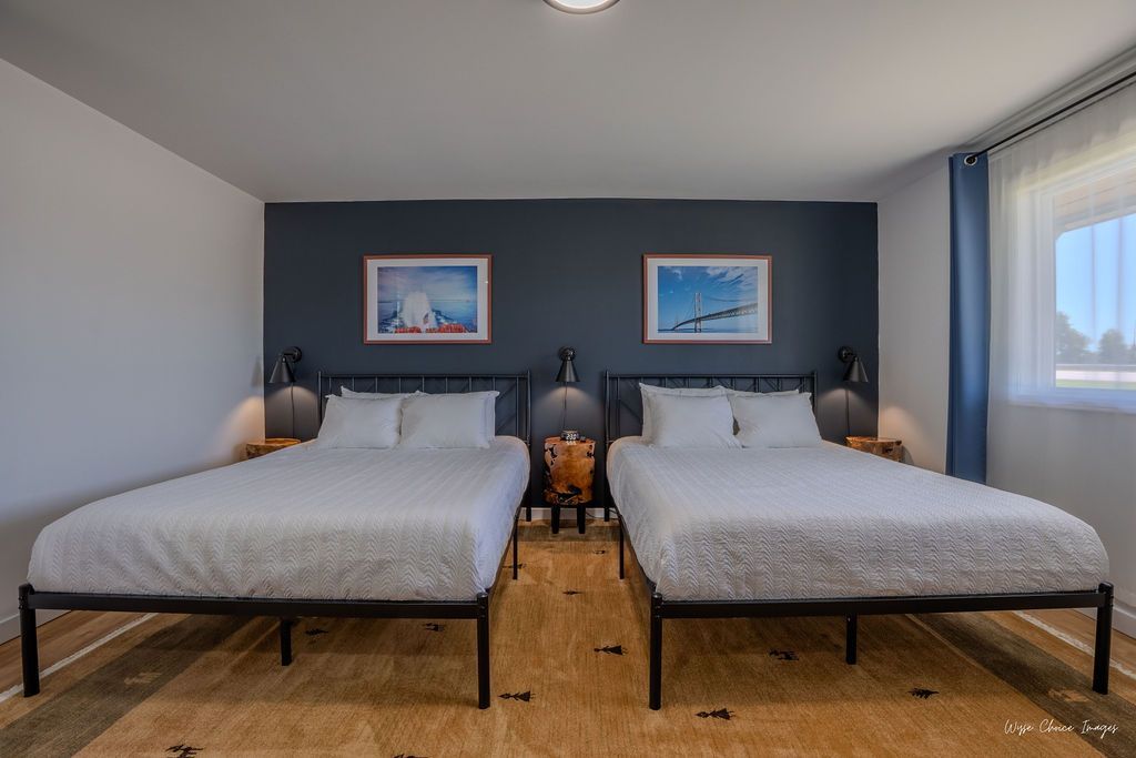Bedroom with two beds against a dark blue wall, with artwork and nightstands. Window on right.
