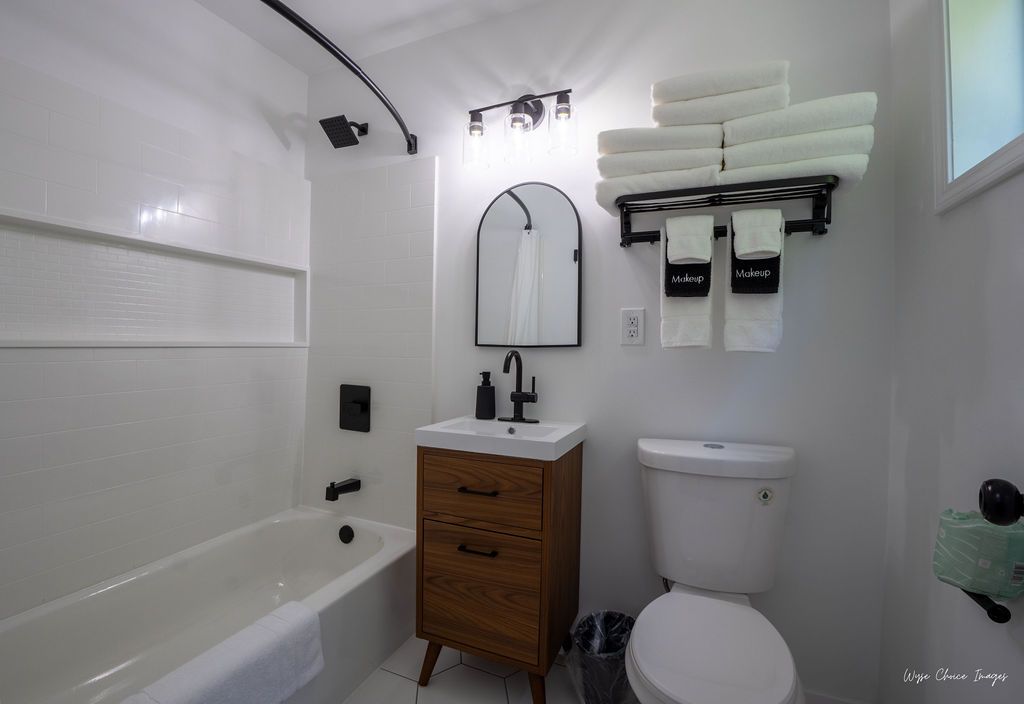Bathroom with white walls, tub, vanity, and toilet. Stacked white towels on a black rack.