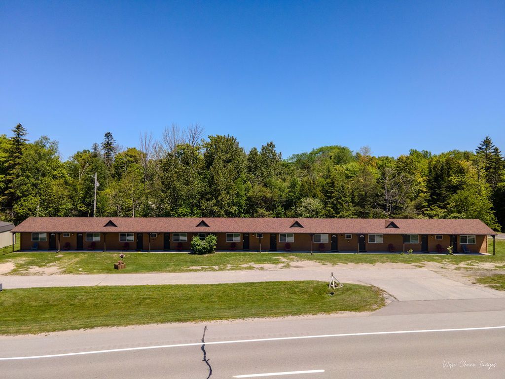 A long, brown-roofed motel with multiple rooms, set against green trees and a blue sky.