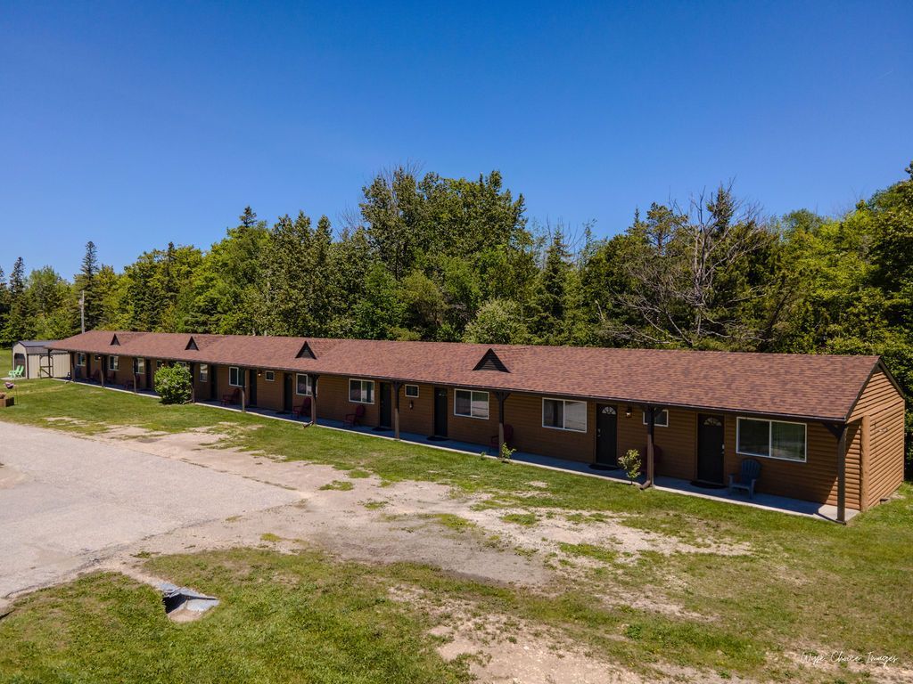 Motel building with brown roof and multiple rooms, set in a grassy area with trees under a blue sky.