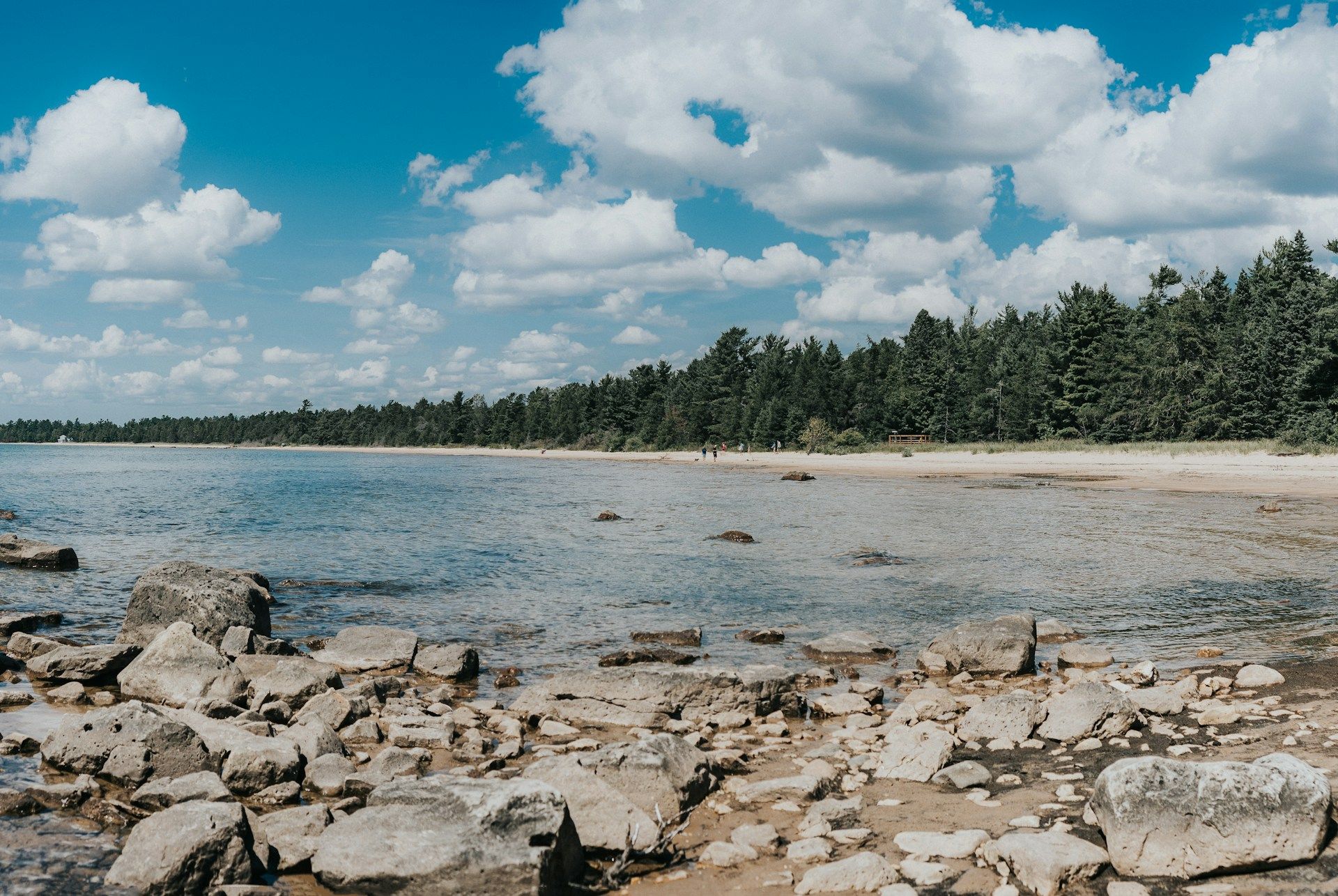 Rocky shoreline meets calm water under a blue sky dotted with white clouds. Trees line the far shore.