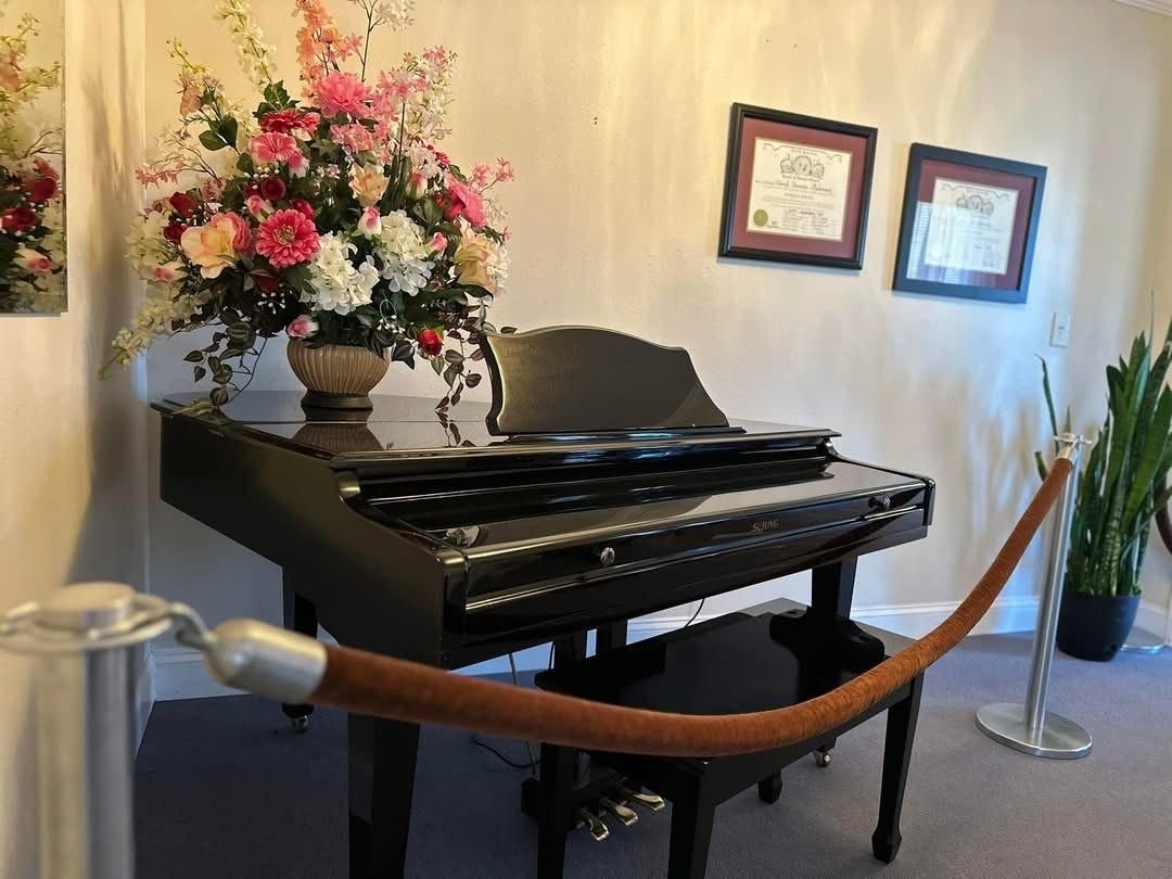 Black grand piano with flower arrangement and roped barrier. Two framed pictures on wall.