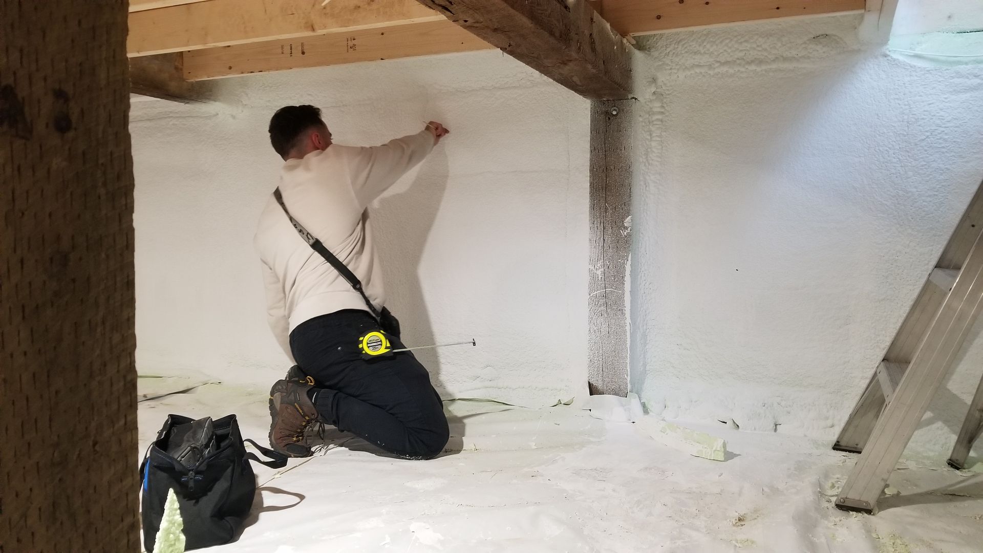 Une personne est agenouillée dans une pièce aux murs isolés en mousse blanche, en train de mesurer un mur. On aperçoit une échelle et un sac noir.