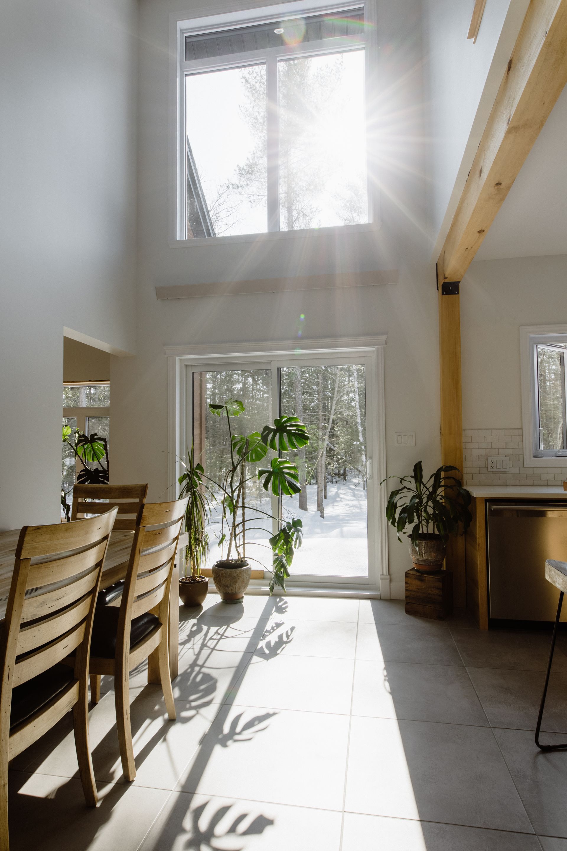 Salle à manger lumineuse et aérée avec de grandes fenêtres, baignée de soleil, et une table en bois avec des chaises. Plantes en pot et vue sur un paysage enneigé.