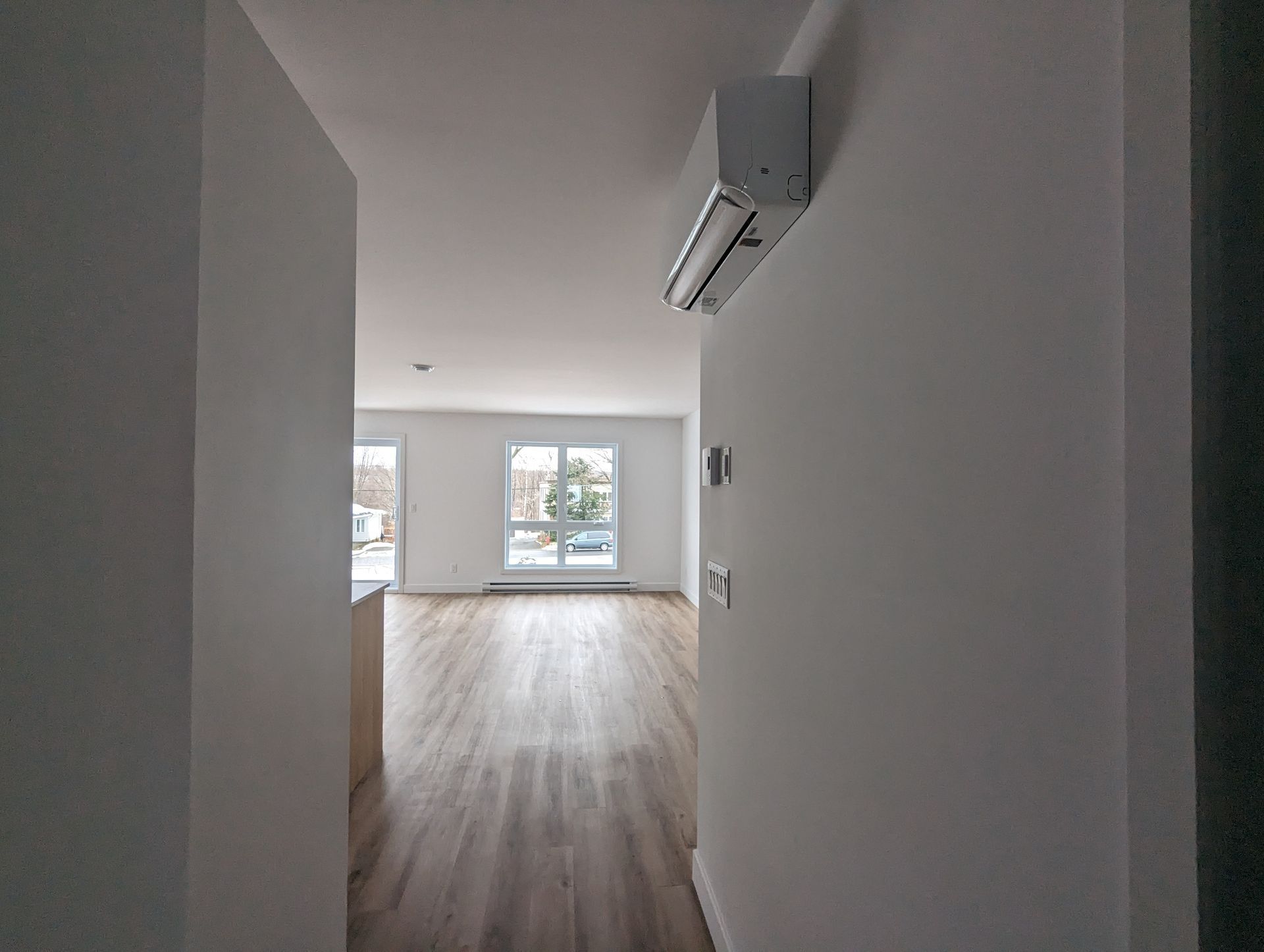 Un couloir mène à une pièce lumineuse avec des fenêtres, un sol imitation bois et un climatiseur mural.