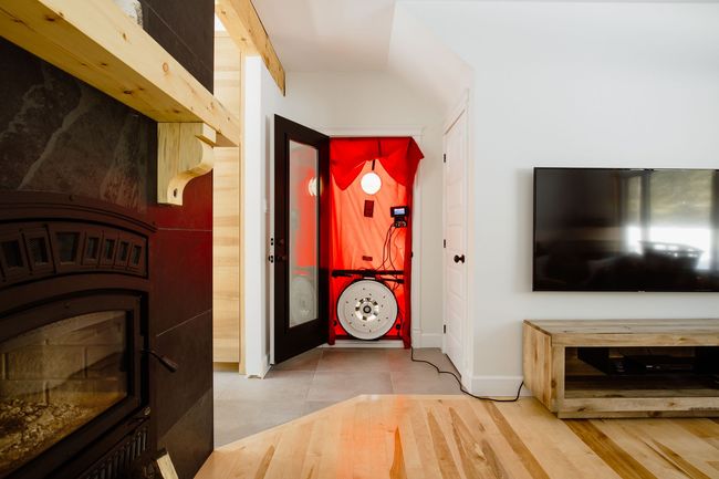Ventilateur dans l'encadrement de la porte lors du test d'étanchéité à l'air ; tissu rouge, porte noire, cheminée et télévision visibles.