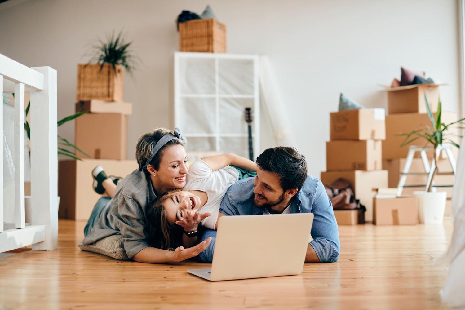 Family lounging on a wooden floor, smiling at a laptop in a bright room with moving boxes.