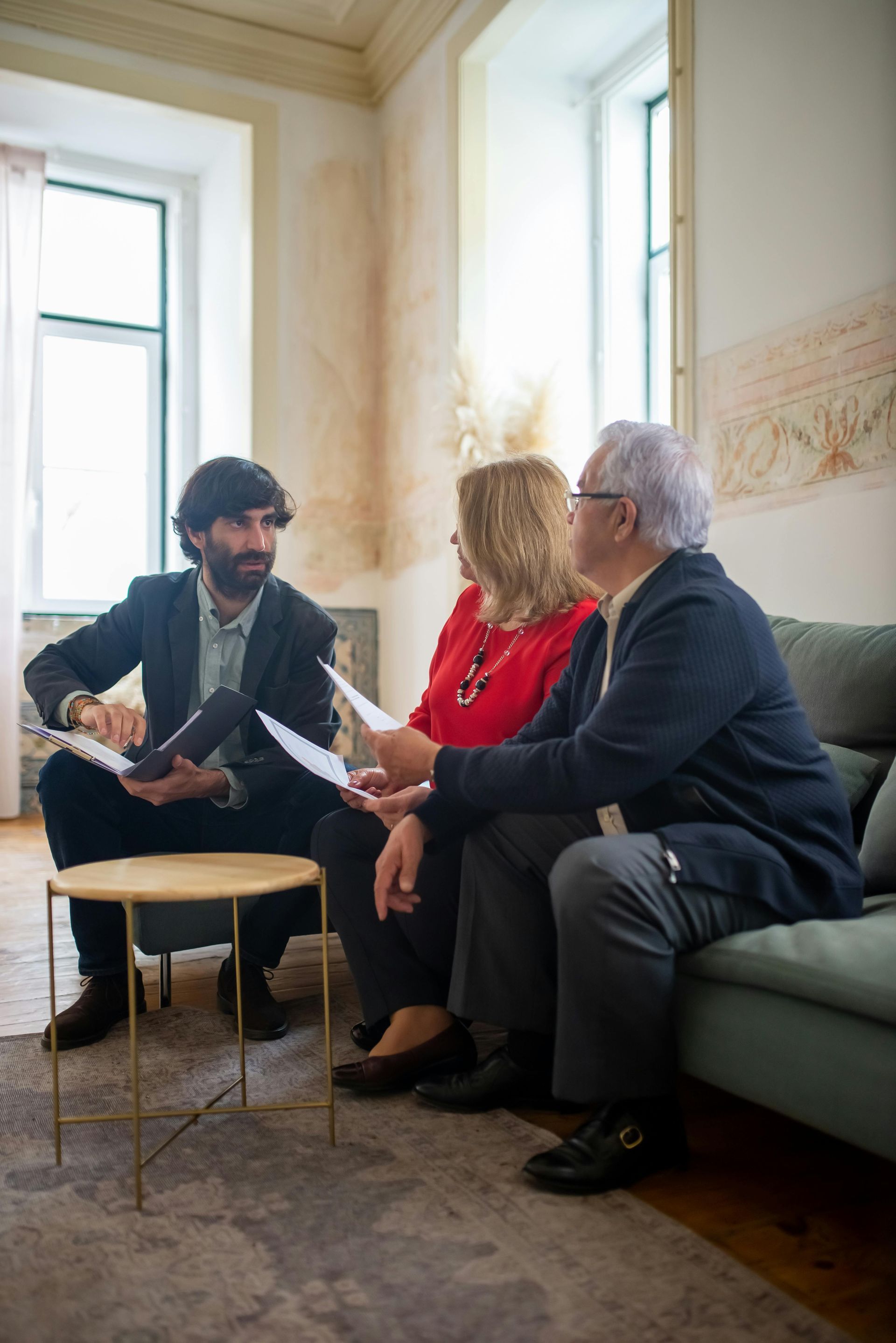 Three people in a bright room discussing papers on a couch and chair