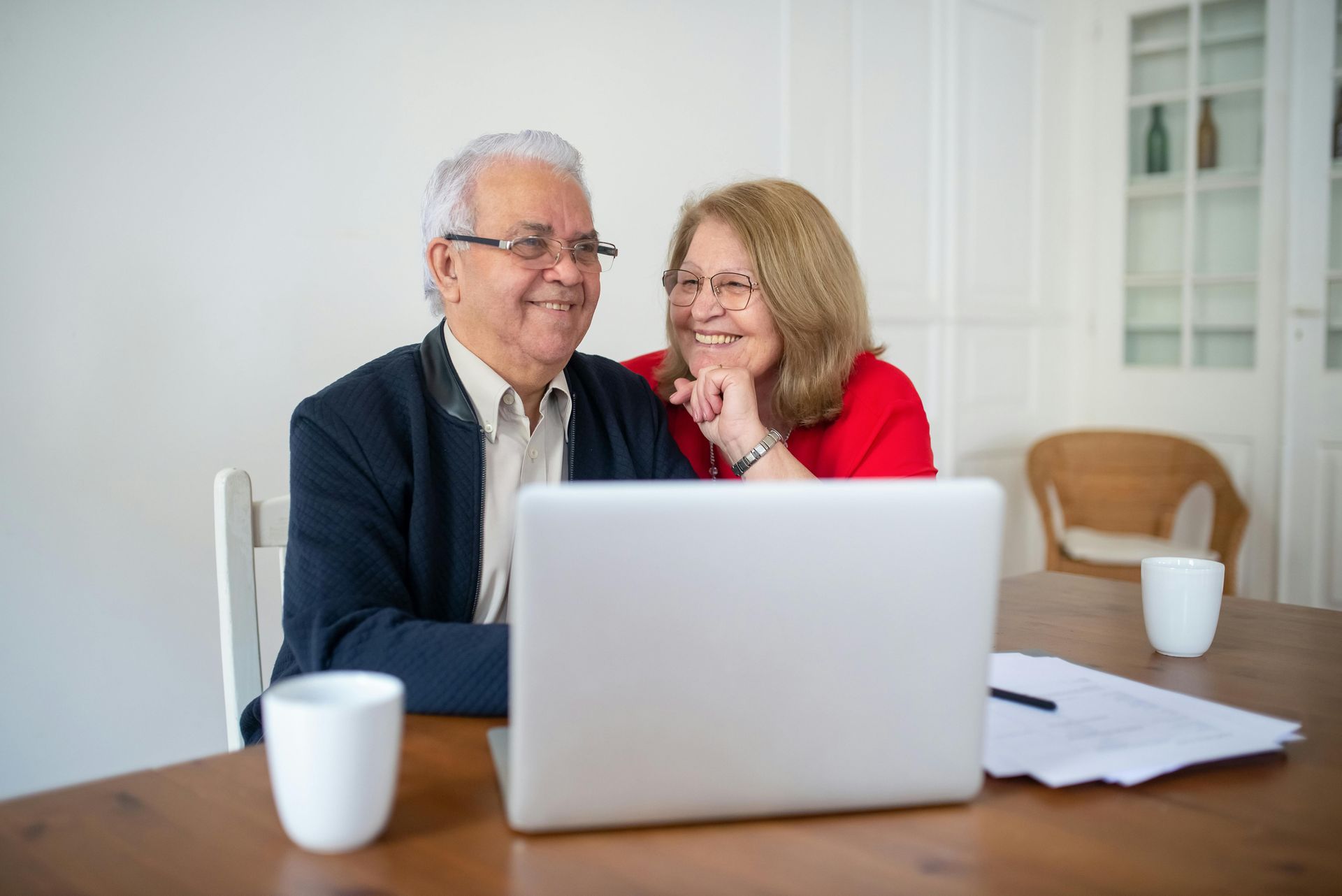 Two people smiling at a laptop on a table, with coffee mugs and papers in a bright office