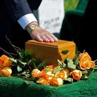 A hand rests on a wooden casket surrounded by orange roses and green foliage.