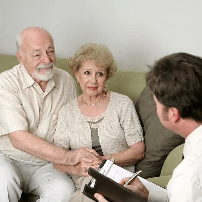 Three people in a living room, seated in discussion with a counselor holding a clipboard.