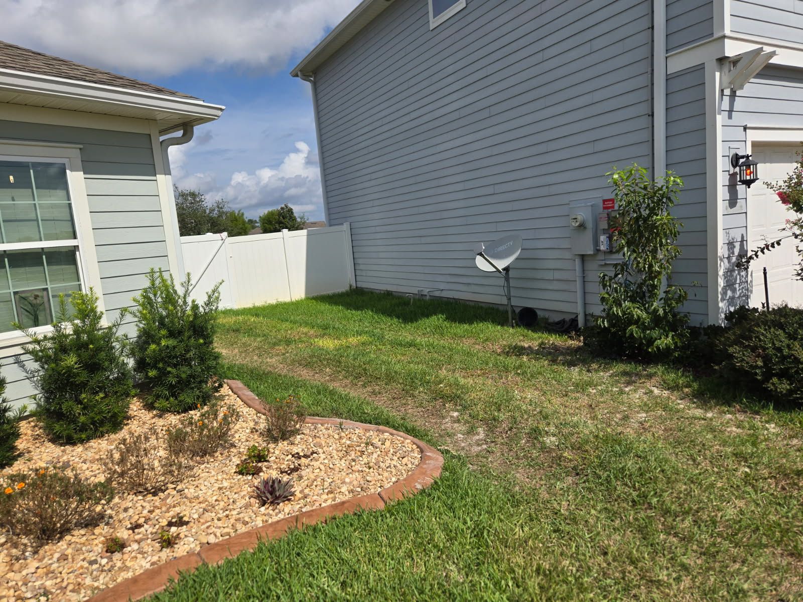 Backyard side yard with grass, white fence, shrubs, and a mulched garden bed beside gray houses